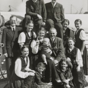 Black-and-white photo of a large group of people posed together outdoors, including several adults and children, with one adult holding a baby at the center.