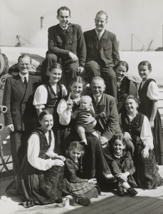 Black-and-white photo of a large group of people posed together outdoors, including several adults and children, with one adult holding a baby at the center.
