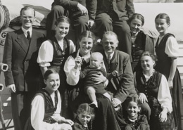 Black-and-white photo of a large group of people posed together outdoors, including several adults and children, with one adult holding a baby at the center.