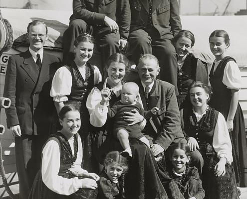 Black-and-white photo of a large group of people posed together outdoors, including several adults and children, with one adult holding a baby at the center.