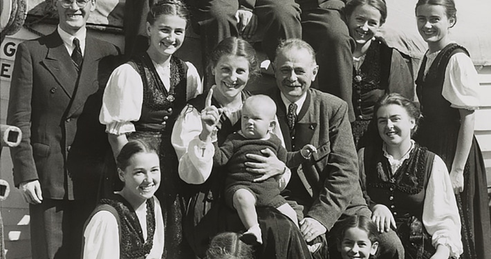 Black-and-white photo of a large group of people posed together outdoors, including several adults and children, with one adult holding a baby at the center.