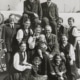 Black-and-white photo of a large group of people posed together outdoors, including several adults and children, with one adult holding a baby at the center.