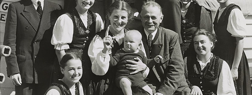 Black-and-white photo of a large group of people posed together outdoors, including several adults and children, with one adult holding a baby at the center.
