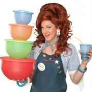 Dixie Longate in Dixie's Tupperware Party, holding colorful Tupperware bowls and a blue cup, with voluminous red hair, wearing a denim shirt and apron against a green background.
