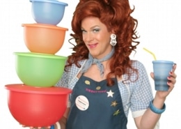Dixie Longate in Dixie's Tupperware Party, holding colorful Tupperware bowls and a blue cup, with voluminous red hair, wearing a denim shirt and apron against a green background.