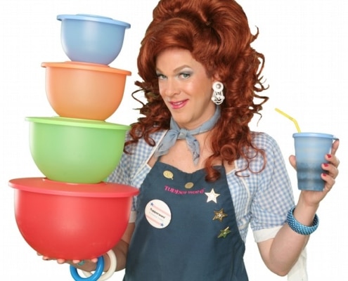 Dixie Longate in Dixie's Tupperware Party, holding colorful Tupperware bowls and a blue cup, with voluminous red hair, wearing a denim shirt and apron against a green background.