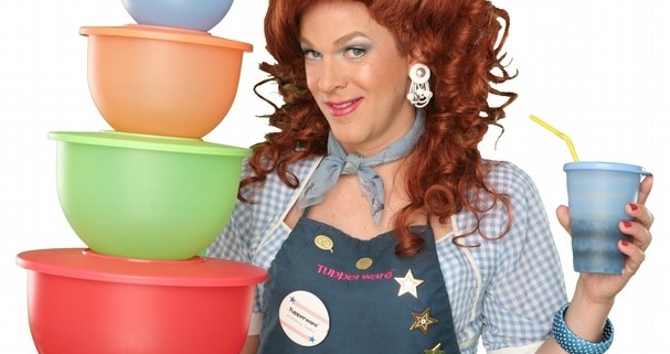 Dixie Longate in Dixie's Tupperware Party, holding colorful Tupperware bowls and a blue cup, with voluminous red hair, wearing a denim shirt and apron against a green background.