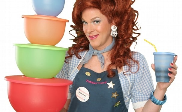 Dixie Longate in Dixie's Tupperware Party, holding colorful Tupperware bowls and a blue cup, with voluminous red hair, wearing a denim shirt and apron against a green background.