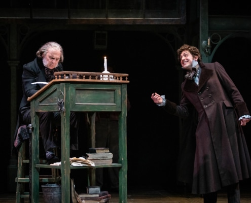 Scrooge sits at a desk while a young man speaks with him in A Christmas Carol