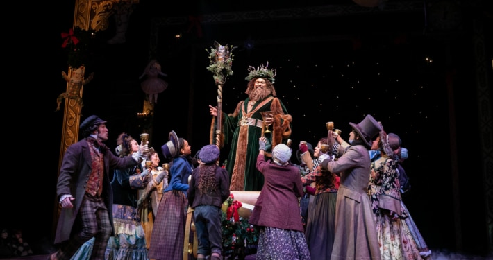 The Ghost of Christmas Present stands on a sleigh above a group of people in A Christmas Carol