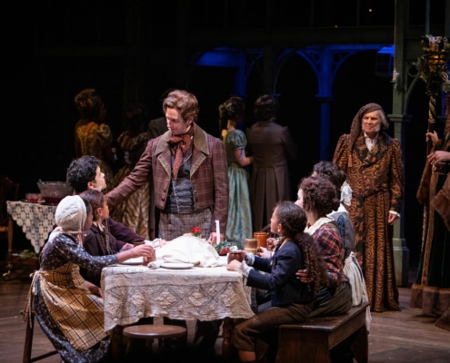 Bob Cratchit stands at his family dinner table while Scrooge watches them in A Christmas Carol