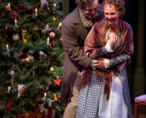 A man embraces his wife from behind, they are both smiling and laughing. The man wears a brown checkered coat, the woman wears a long blue dress and a pink shawl. There is a large Christmas tree in the background.