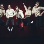 Five men in white shirts and black knickers are photographed in mid-air while jumping in front of a black background.