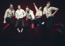 Five men in white shirts and black knickers are photographed in mid-air while jumping in front of a black background.