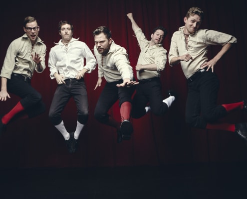 Five men in white shirts and black knickers are photographed in mid-air while jumping in front of a black background.