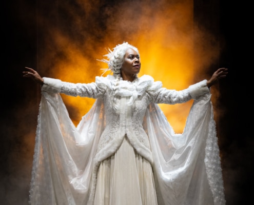A woman, The Ghost of Christmas Past, regally spreads out her arms, she looks to the right. She is wearing a long, intricate, white dress lined with white fur, a long white cape is connected to her wrists and spreads out behind her.