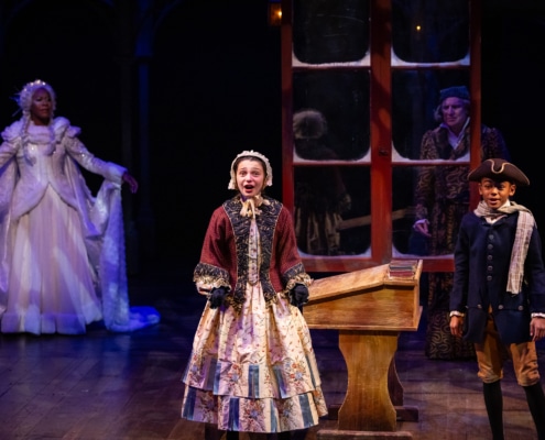 A young boy and girl dressed in Victorian grab, stand and sing next to an old wooden desk. Ebenezer Scrooge dressed in his robe and night cap looks at his child self through a window. The Ghost of Christmas Past observes from the side.