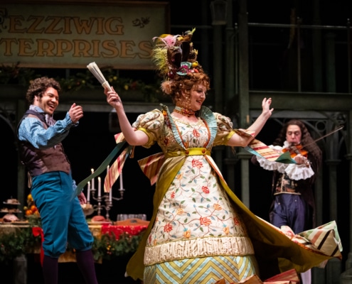 A man wearing a brown vest and bright blue pants grins and the woman in the center of the room. The woman is twirling in a brightly colored and patterned Victorian dress, she is holding a fan, her hair is styled up with feathers in her hair. There is a man playing the fiddle in the background.