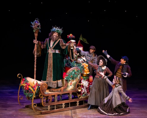 The Ghost of Christmas Present wears a green velvet robe and a crown, holding a wooden staff and a gold goblet. He is standing on a gilded sleigh that carries food and presents. Four young people wearing Victorian garb grin and gesture towards him.