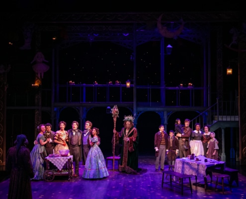 Ebenezer Scrooge faces away from the camera. He is looking at The Ghost of Christmas Present who stands between two groups of smiling singing people. The group on the left is a group of young adults wearing Victorian garb. The group on the right is a family with young children the father holds one child in his arms.