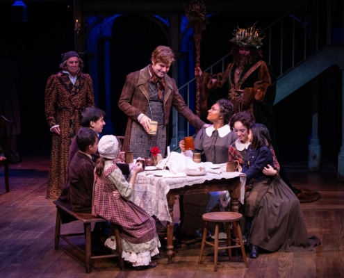 Ebenezer Scrooge and The Ghost of Christmas Present look on at the Cratchit family. The family is sitting around a simple table, the father is standing at the head of the table making a toast, three are three people on the two benches lining the table.