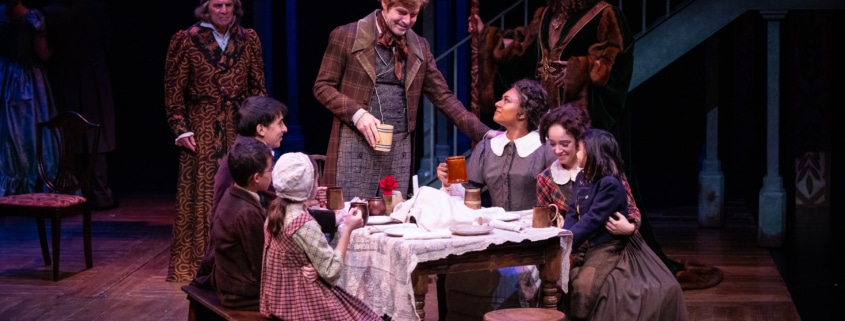 Ebenezer Scrooge and The Ghost of Christmas Present look on at the Cratchit family. The family is sitting around a simple table, the father is standing at the head of the table making a toast, three are three people on the two benches lining the table.