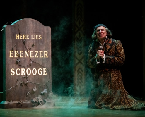 A man in a long robe and a gray night cap kneels in front of his gravestone, his hands clasped. The gravestone reads Here Lies Ebenezer Scrooge.