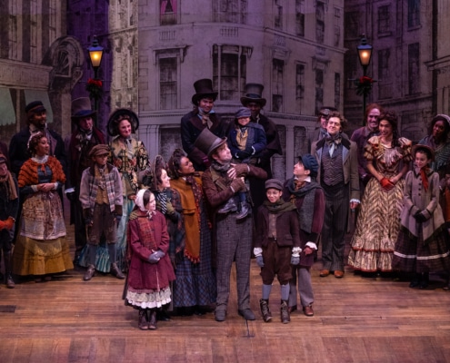 A family stands in the center of the stage; they smile at each other. The father holds his son, Tiny Tim on his shoulder. Other people dressed in Victorian garb look on at the family smiling.