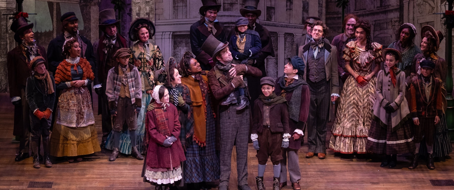 A family stands in the center of the stage; they smile at each other. The father holds his son, Tiny Tim on his shoulder. Other people dressed in Victorian garb look on at the family smiling.