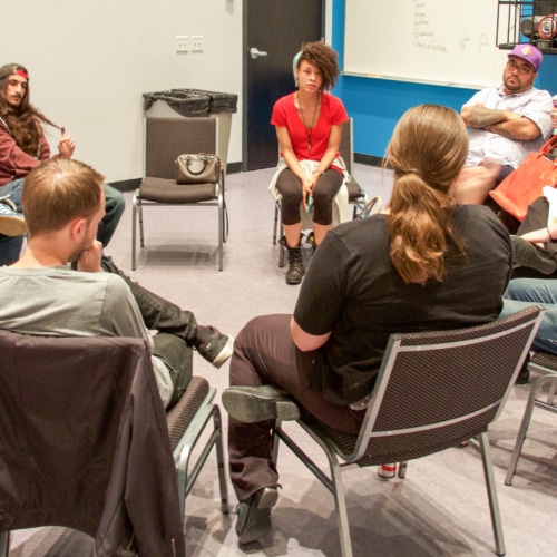 A group of actors sit in a circle in a studio, some holding notepads and paper