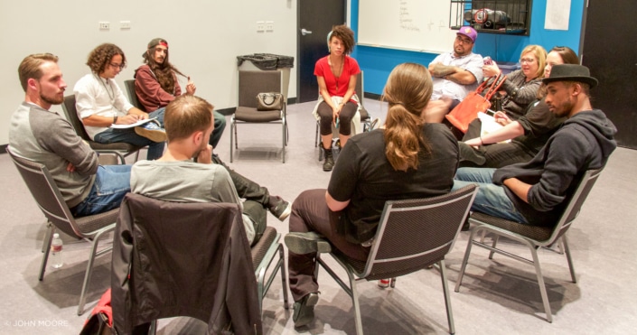 A group of actors sit in a circle in a studio, some holding notepads and paper