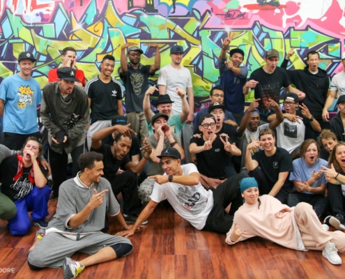 A large troupe of dancers poses for a group shot in front of a wall of graffiti art