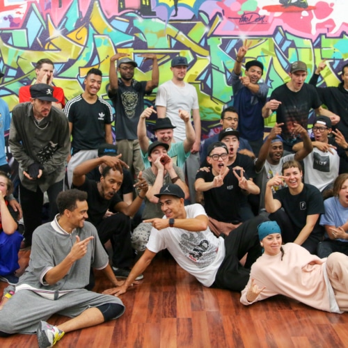 A large troupe of dancers poses for a group shot in front of a wall of graffiti art