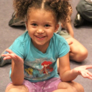 Younger girl sitting with her legs crossed on the floorl in a teal little mermaid shirt is smiling to the camera and shrugging to the cameraman