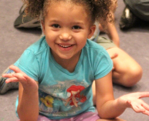 Younger girl sitting with her legs crossed on the floorl in a teal little mermaid shirt is smiling to the camera and shrugging to the cameraman
