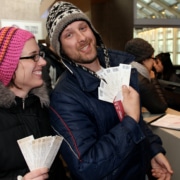 Close-up of a man and woman at a ticket window. Both are dressed in warm winter gear. The man holds a stack of white tickets toward the camera, and the woman holds a similar set. In the background, a staff member and other patrons are visible near a desk with a binder.