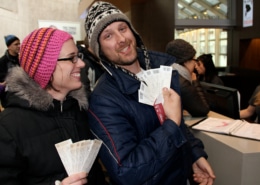 Close-up of a man and woman at a ticket window. Both are dressed in warm winter gear. The man holds a stack of white tickets toward the camera, and the woman holds a similar set. In the background, a staff member and other patrons are visible near a desk with a binder.