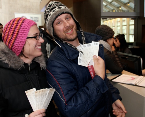 Donate your unused tickets Close-up of a man and woman at a ticket window. Both are dressed in warm winter gear. The man holds a stack of white tickets toward the camera, and the woman holds a similar set. In the background, a staff member and other patrons are visible near a desk with a binder.