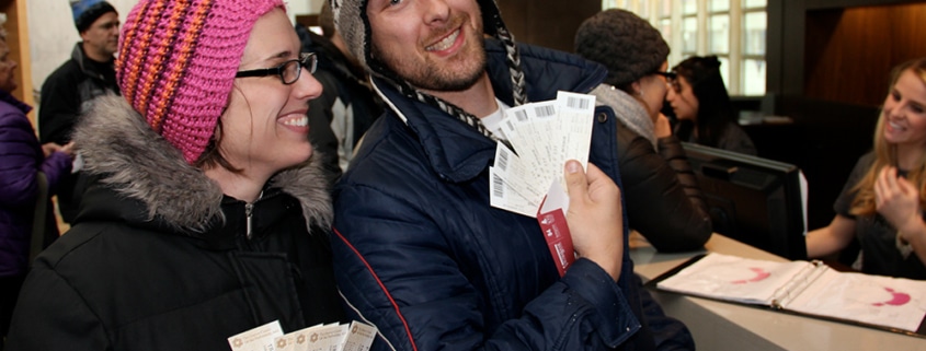 Close-up of a man and woman at a ticket window. Both are dressed in warm winter gear. The man holds a stack of white tickets toward the camera, and the woman holds a similar set. In the background, a staff member and other patrons are visible near a desk with a binder.