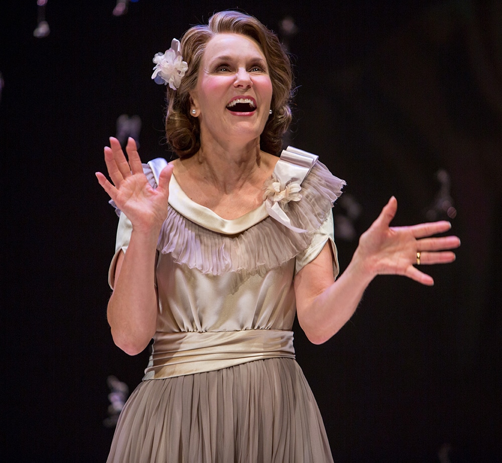 Woman in beige dress with a flower in her hair smiles in awe outwards as small floating glass trinkets are blurred behind her