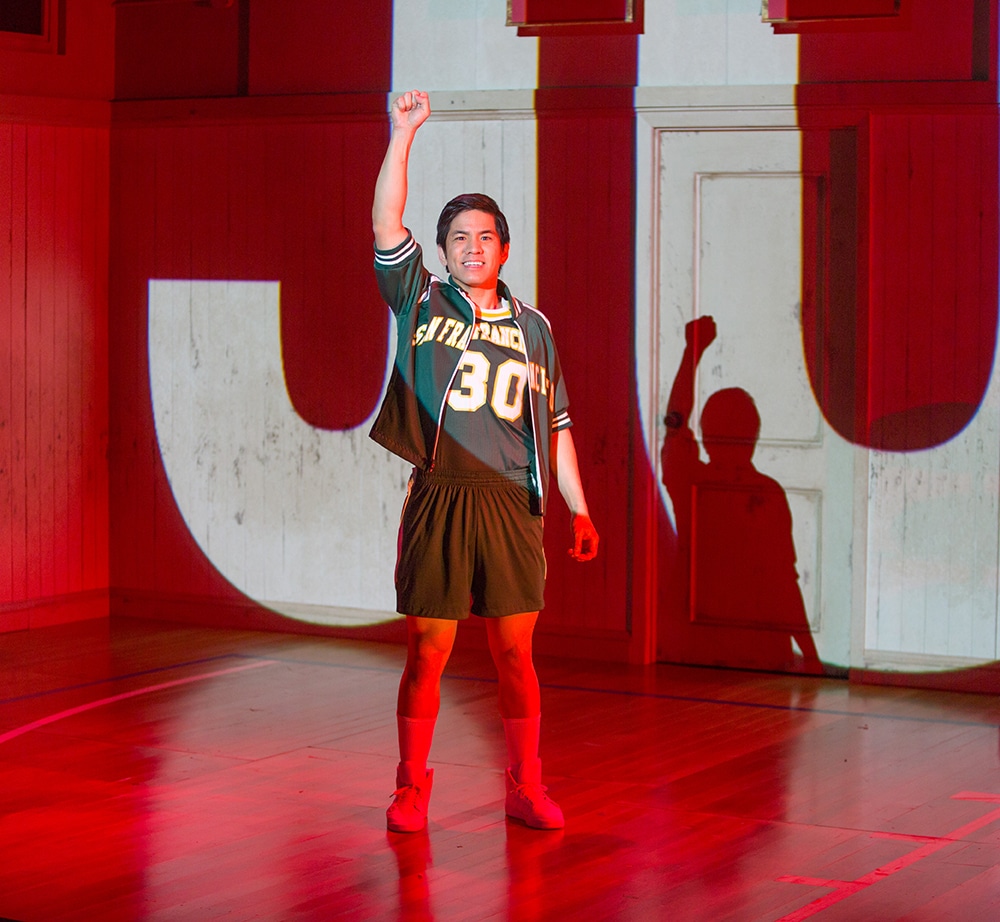 Man in a forest green basketball uniform and varsity jacket stands triumphantly smiling with his right fist in the air