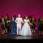 Grandview High School students take a bow during a stage production, wearing elaborate period costumes against a vibrant pink backdrop.