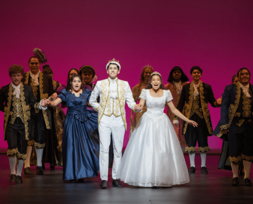Grandview High School students take a bow during a stage production, wearing elaborate period costumes against a vibrant pink backdrop.