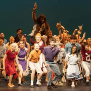Denver School of the Arts students perform a lively ensemble number on stage, wearing colorful costumes and holding props during a theatrical production.