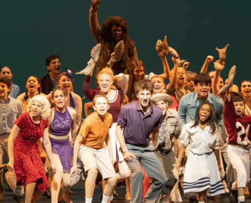 Denver School of the Arts students perform a lively ensemble number on stage, wearing colorful costumes and holding props during a theatrical production.