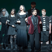 Vista Ridge High School students perform a scene from a stage production, wearing dark, character-themed costumes under dramatic theatrical lighting.