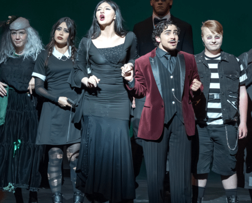 Vista Ridge High School students perform a scene from a stage production, wearing dark, character-themed costumes under dramatic theatrical lighting.