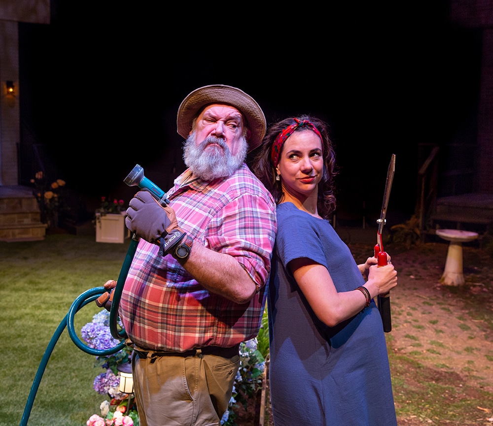 Man in a red plaid shirt and fisherman hat holds a hose standing back-to-back with woman in blue holding hedge shears