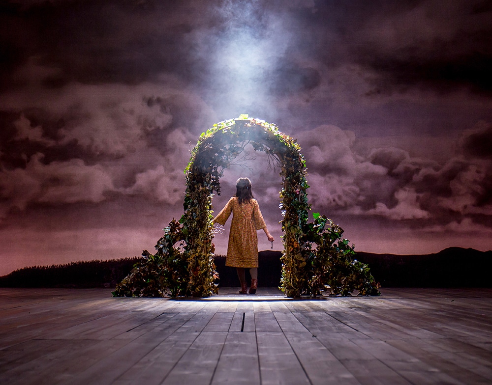 Young girl in early 20th century peasant dress walks upstage through an arch made up of greenery with a moody cloud backdrop