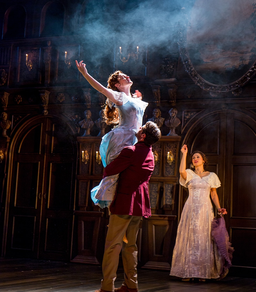 Man lifts a woman dressed in early 20th century royal dress as she stretches upwards while another woman watches from behind